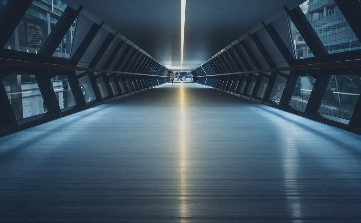 A covered pedestrian passage between office towers in a financial district, lit from the far end, with the walkway narrowing toward a single controlled opening.
