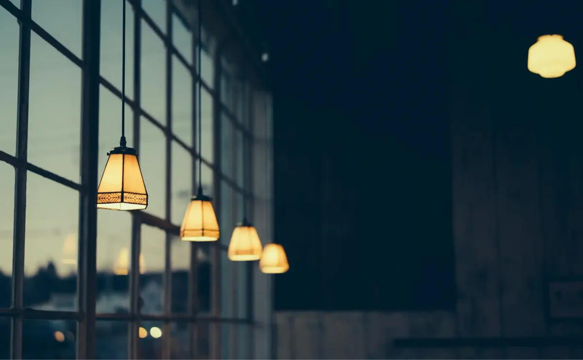 A receding row of pendant lamps along a dark window wall, the nearest one sharp and lit and each one behind it fading further into shadow.